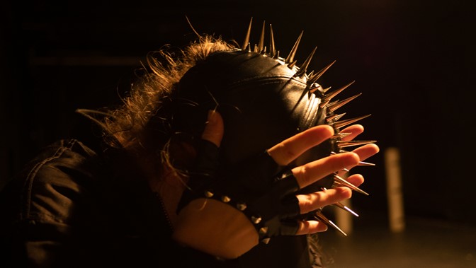 A performers in a spiked headpiece holds their head in their hands
